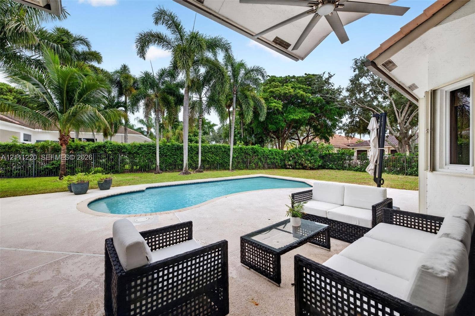 134 Dockside Terrace, Weston, FL 33327 Covered patio featuring an integrated ceiling fan, concrete deck, kidney-shaped swimming pool, lush perimeter hedge, and mature palm trees