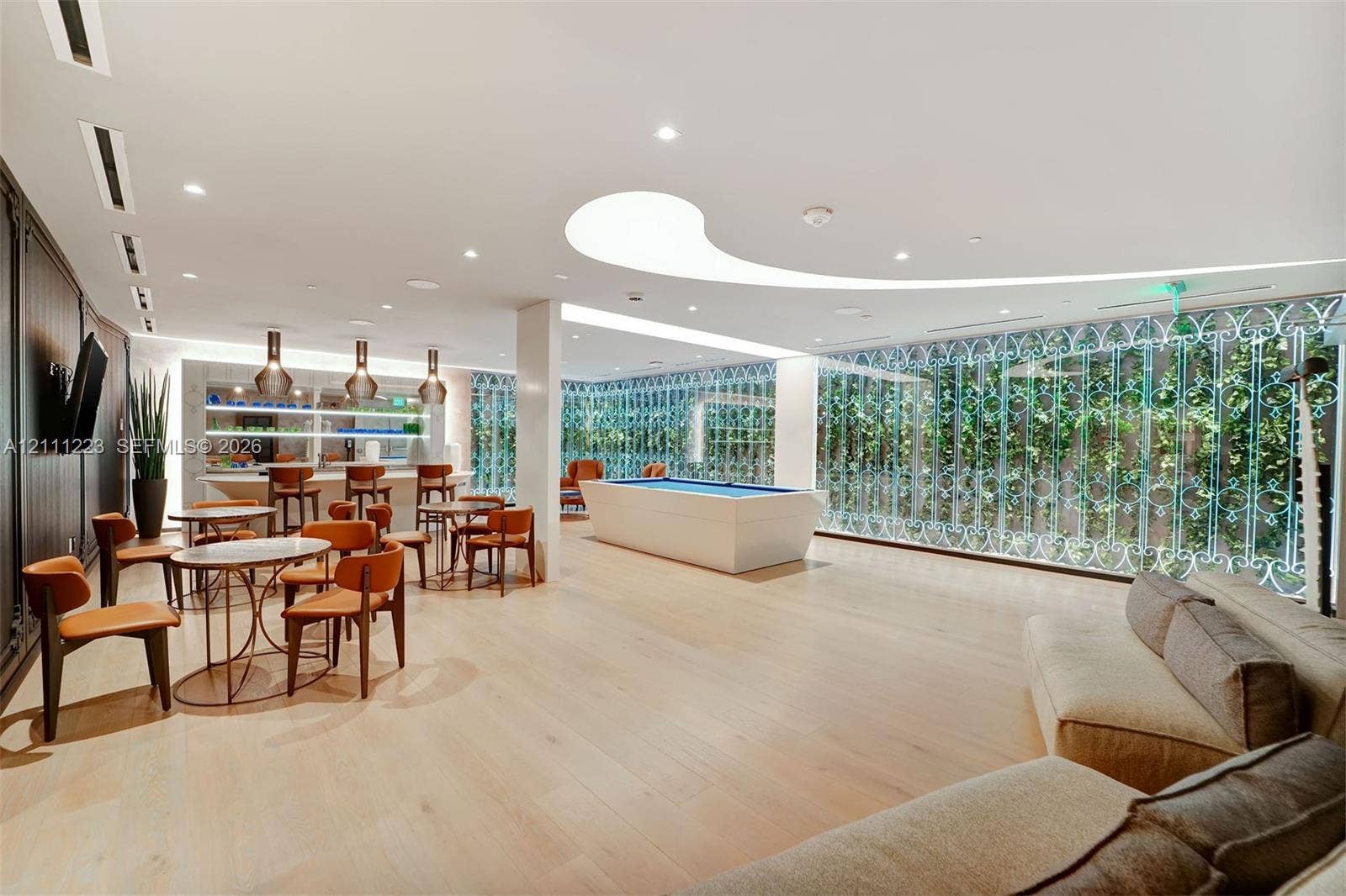 Large common area featuring light-toned wood-finish flooring, a curved ceiling cove light fixture, built-in bar with pendant lighting, and expansive floor-to-ceiling lattice screen windows