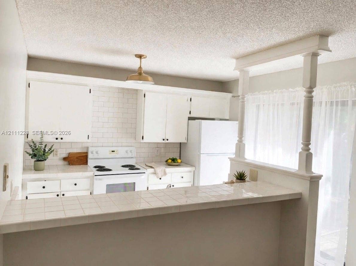 Kitchen featuring white cabinetry, subway tile backsplash, ceramic tile countertop, pass-through breakfast bar with decorative columns, and metallic pendant lighting