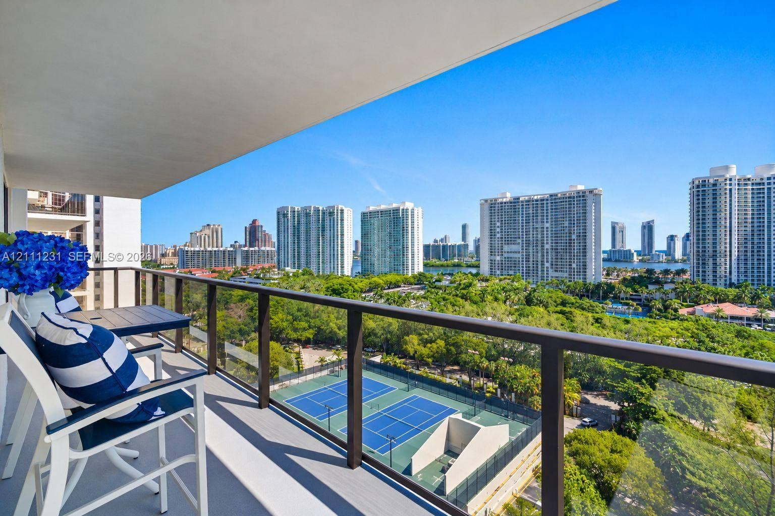 Covered private balcony featuring a glass and dark metal railing overlooking dual tennis courts and surrounding urban landscape