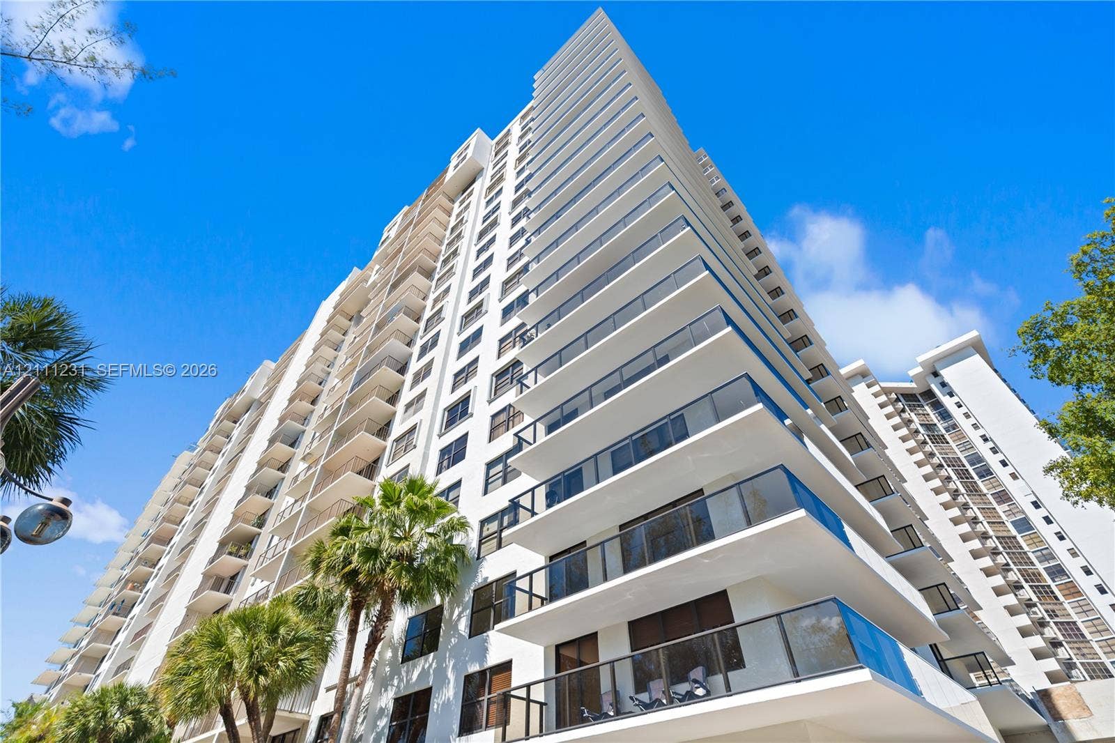 High-rise residential exterior featuring white concrete facade, stacked private balconies, glass railings, and expansive windows