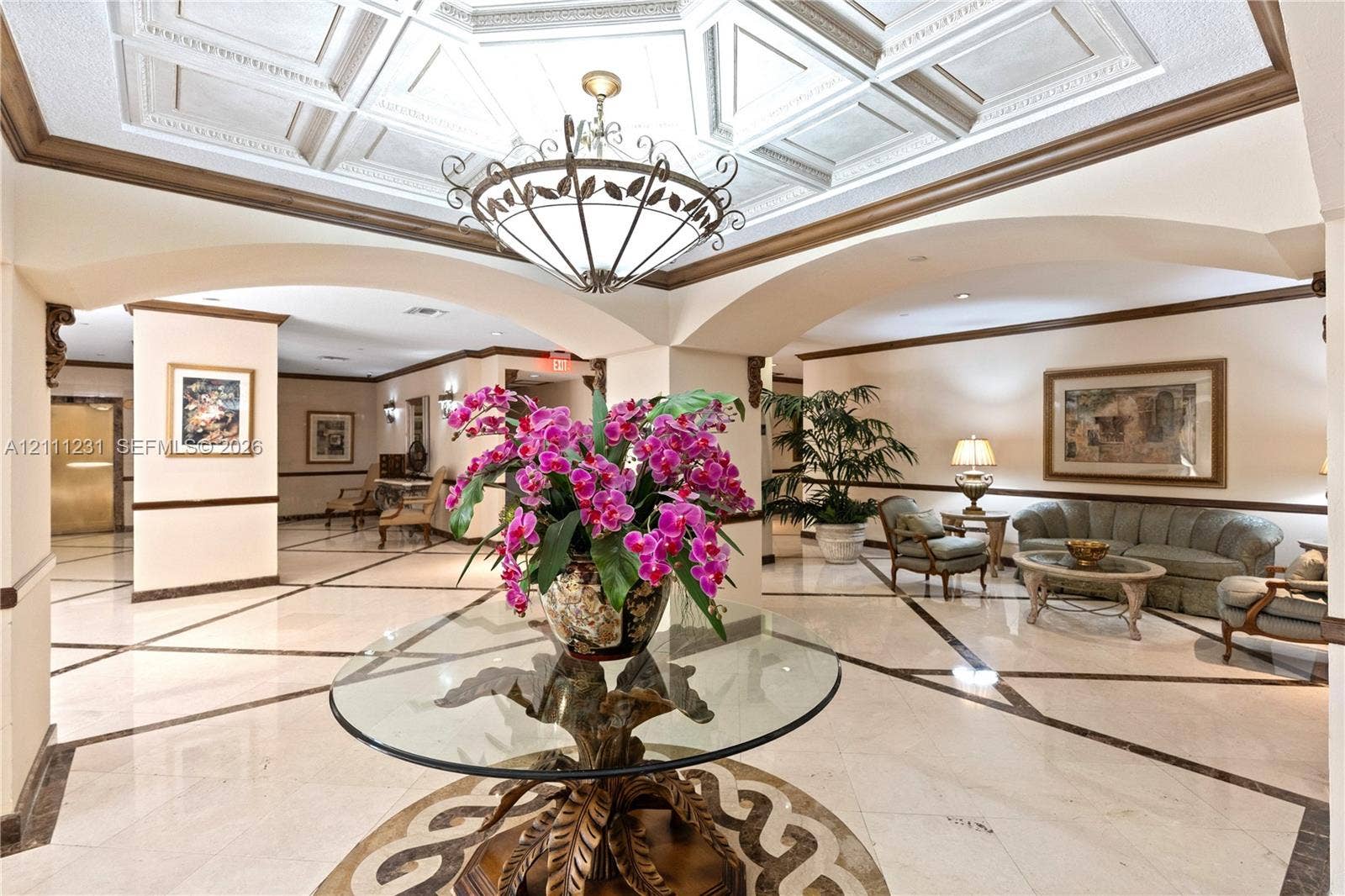 Grand foyer featuring intricate coffered ceiling detailing, stone-finish flooring with decorative inlay, arched architectural openings, and a prominent multi-arm central chandelier