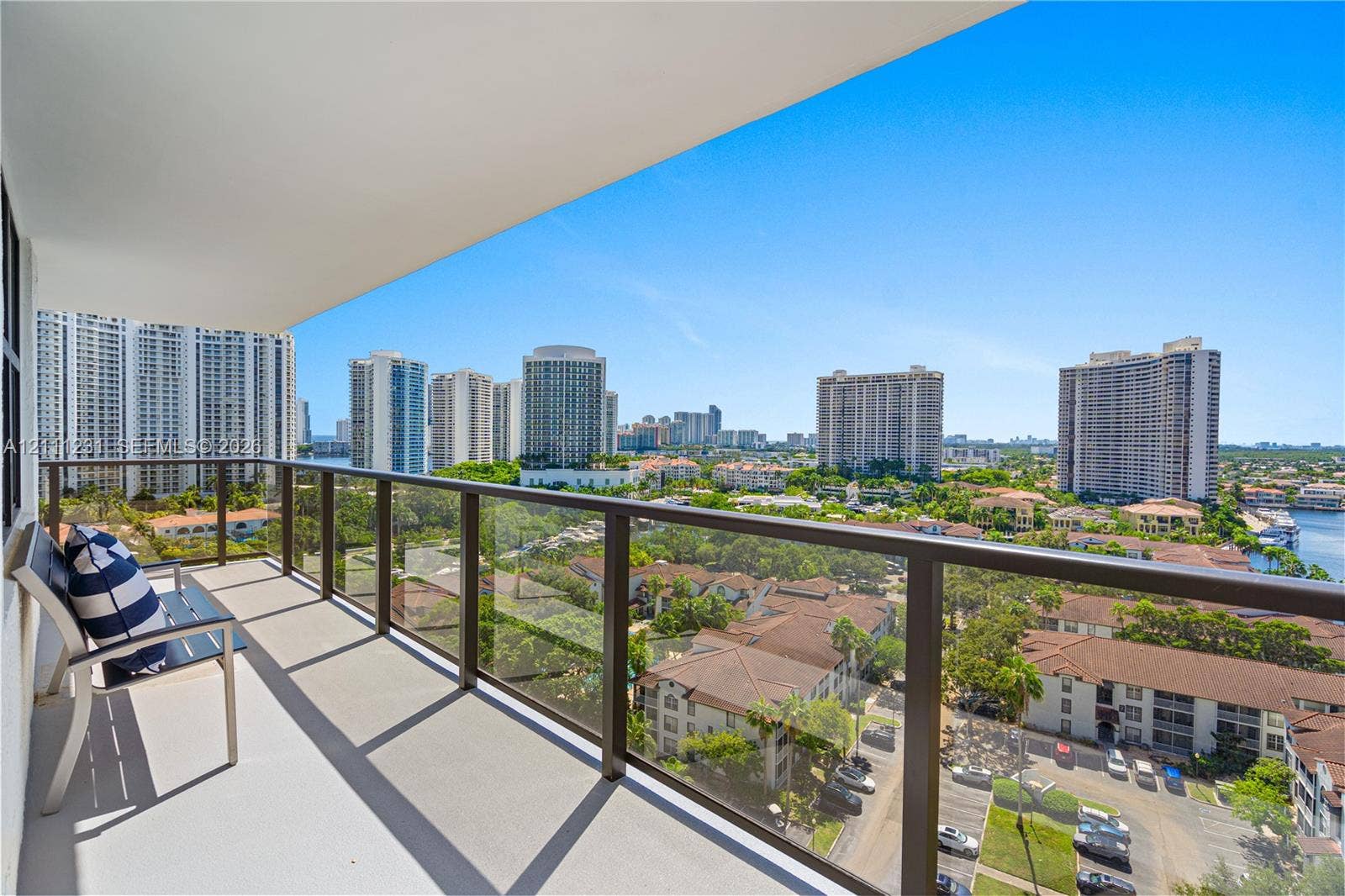 Covered balcony featuring concrete flooring and a dark-finish metal railing with glass panels, providing an expansive perspective of the surrounding skyline and urban landscape
