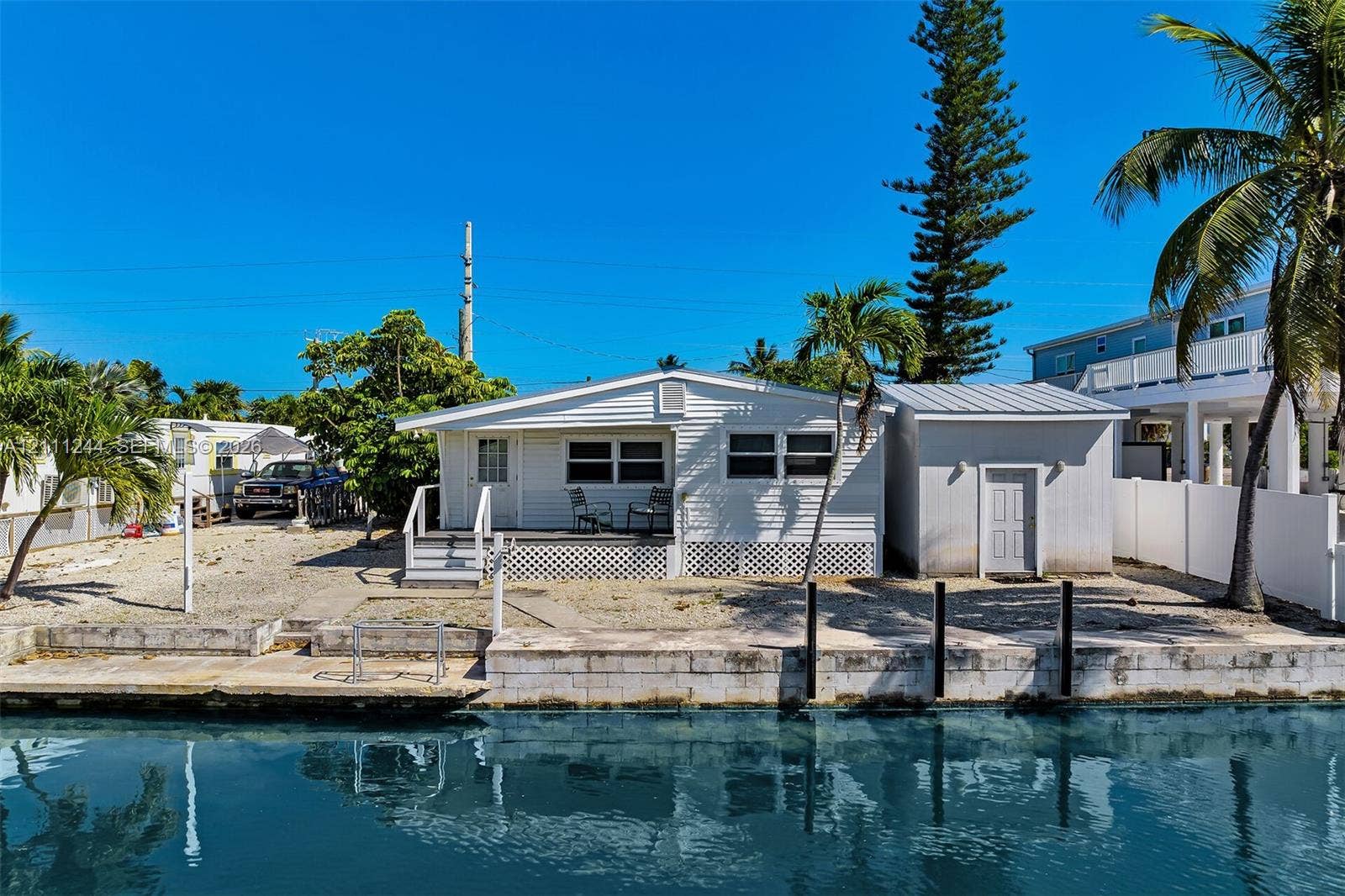 Waterfront exterior featuring light-colored horizontal siding, a raised deck with lattice skirting, an attached utility structure, and concrete seawall bulkhead