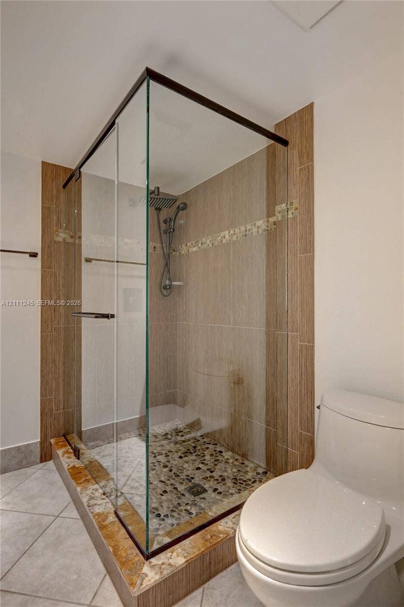 6800 Cypress Road, #506, Plantation, FL 33317 Walk-in glass-enclosed shower featuring a river rock floor, multi-head shower system, decorative tile accents, and stone-tiled shower curb