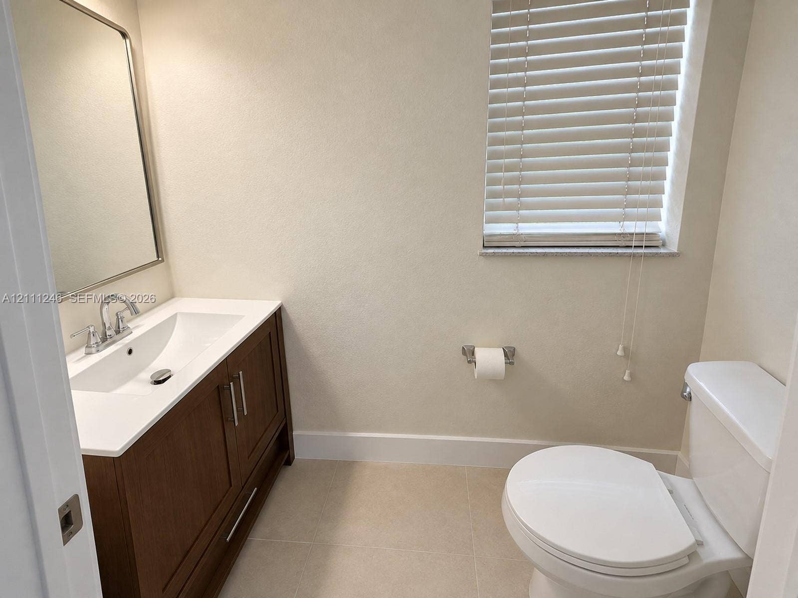 Vanity featuring a dark wood-finish cabinet, integrated white sink, polished chrome faucet, and rectangular framed mirror, complemented by neutral tile flooring and window blinds