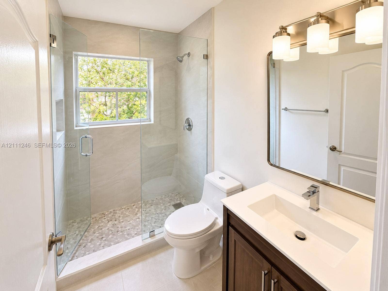 Walk-in shower with a glass enclosure, pebble-tile flooring, and built-in wall niche, featuring a wood-finish vanity with a rectangular sink and a multi-light fixture above the mirror