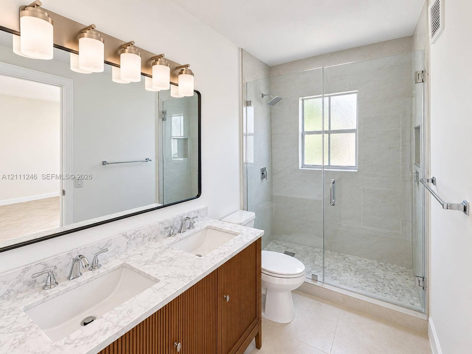 Dual-sink vanity with fluted cabinetry and stone-finish countertop, frameless glass-enclosed shower featuring tiled interior, and oversized wall-mounted mirror with integrated five-light fixture