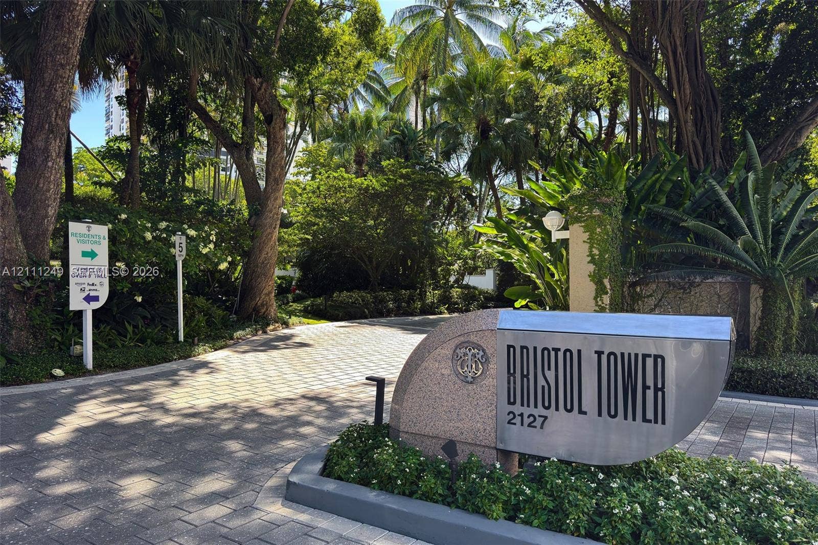 Gated property entrance featuring a masonry monument sign, paver driveway, and mature tropical landscaping