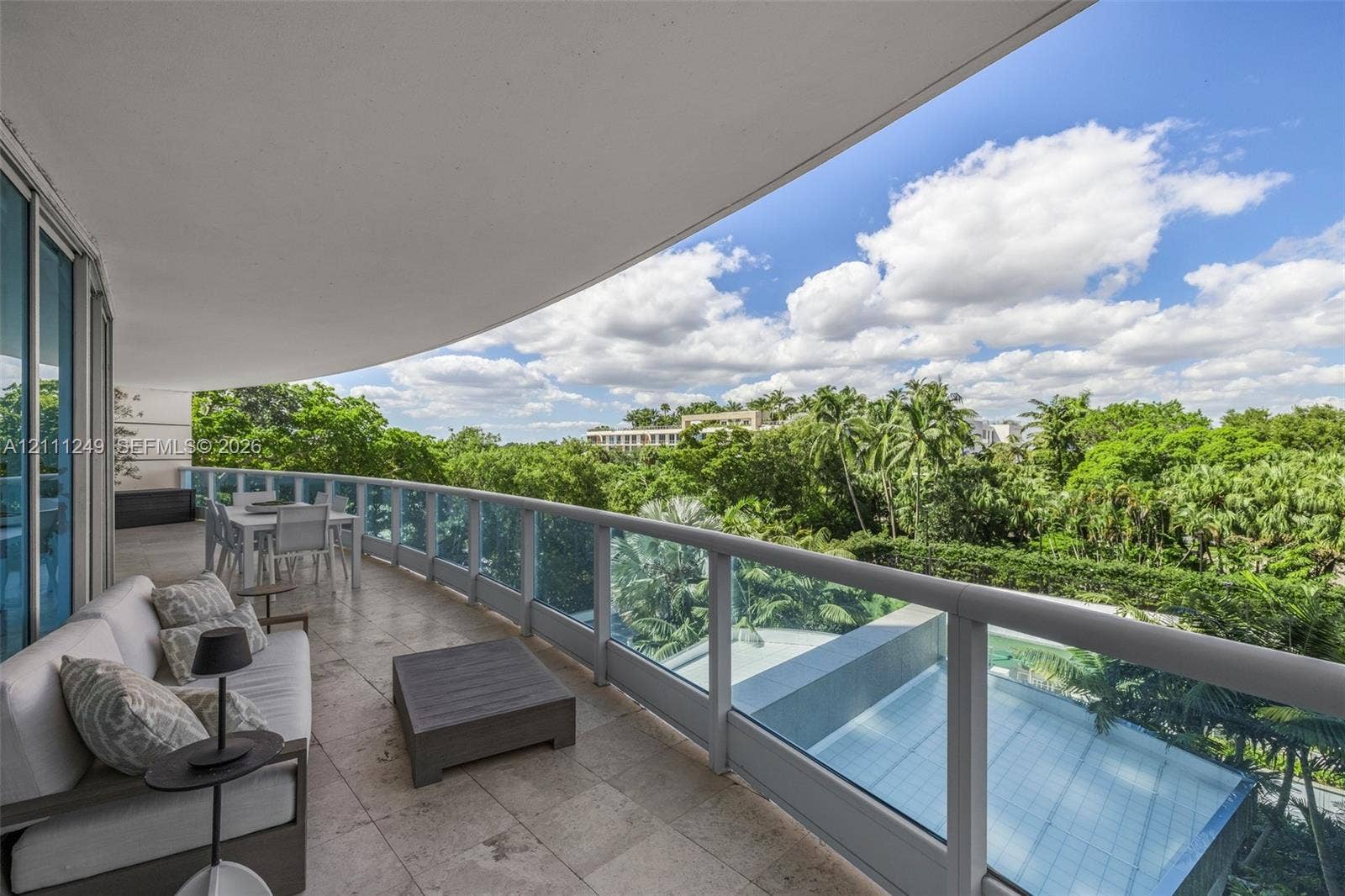 Expansive covered balcony featuring stone flooring, a glass-panel railing, and direct exposure to mature palm trees and landscaping
