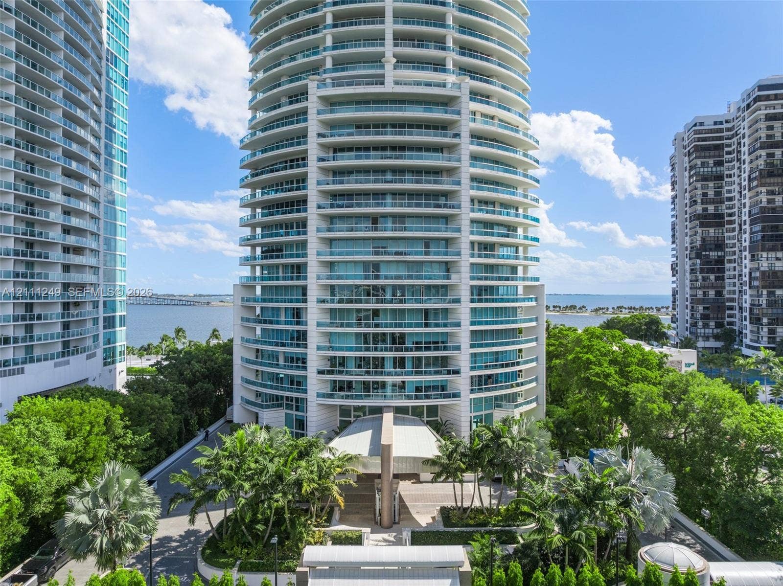 High-rise residential tower featuring a glass and concrete facade, wrap-around balconies with glass railings, a covered porte-cochre entry, and landscaped grounds