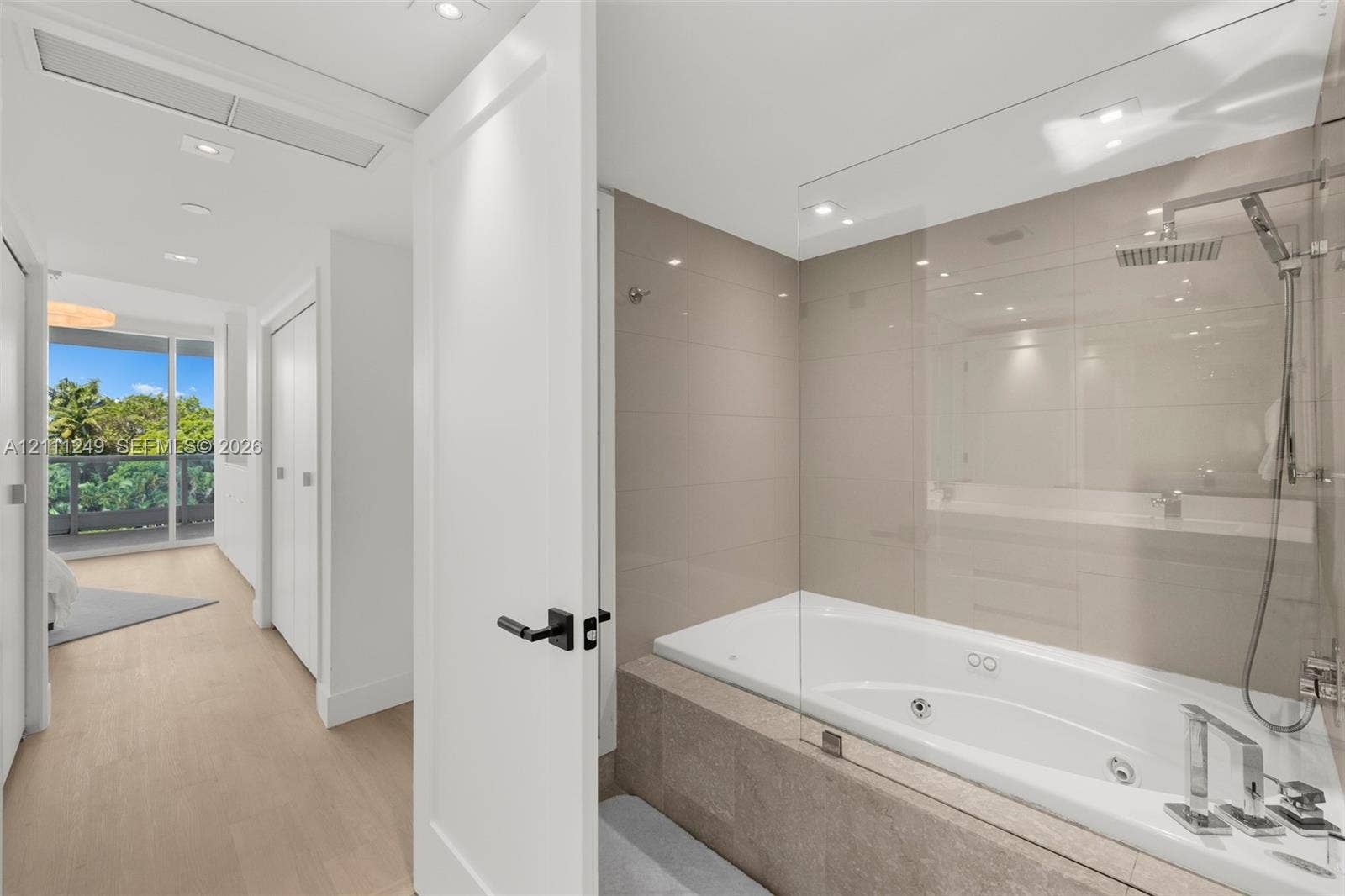 Jetted soaking tub with a glass partition, large-format wall tile, polished metal plumbing fixtures, and wood-finish flooring