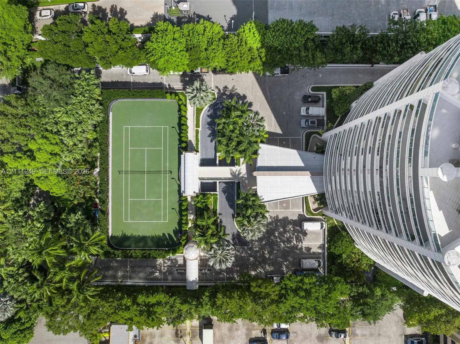 Aerial view of the high-rise structure featuring an on-site tennis court, paved parking area, and mature landscaping
