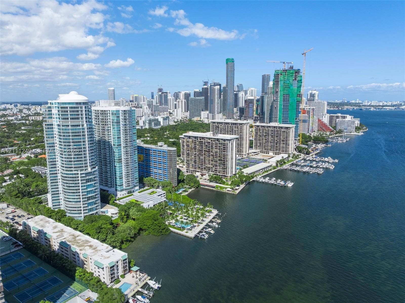 Aerial perspective of a high-density waterfront environment featuring multi-story residential towers, a deep-water marina, hard-surface tennis courts, and expansive bay frontage
