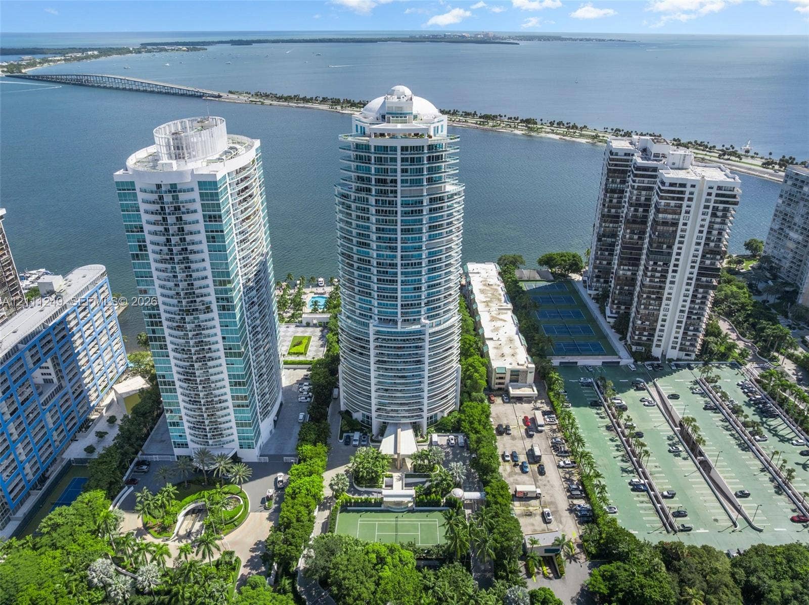 High-rise residential tower featuring glass-paneled balconies, direct coastal proximity, paved tennis courts, and landscaped grounds