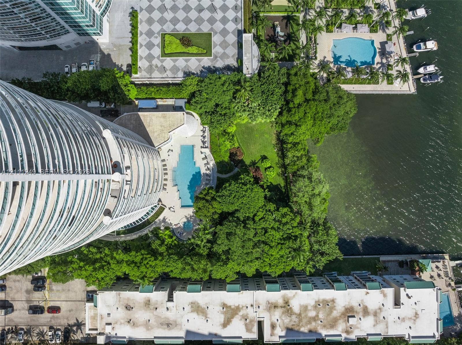 Aerial perspective featuring a high-rise structure, an outdoor swimming pool, landscaped lawn, mature trees, and proximity to the waterfront