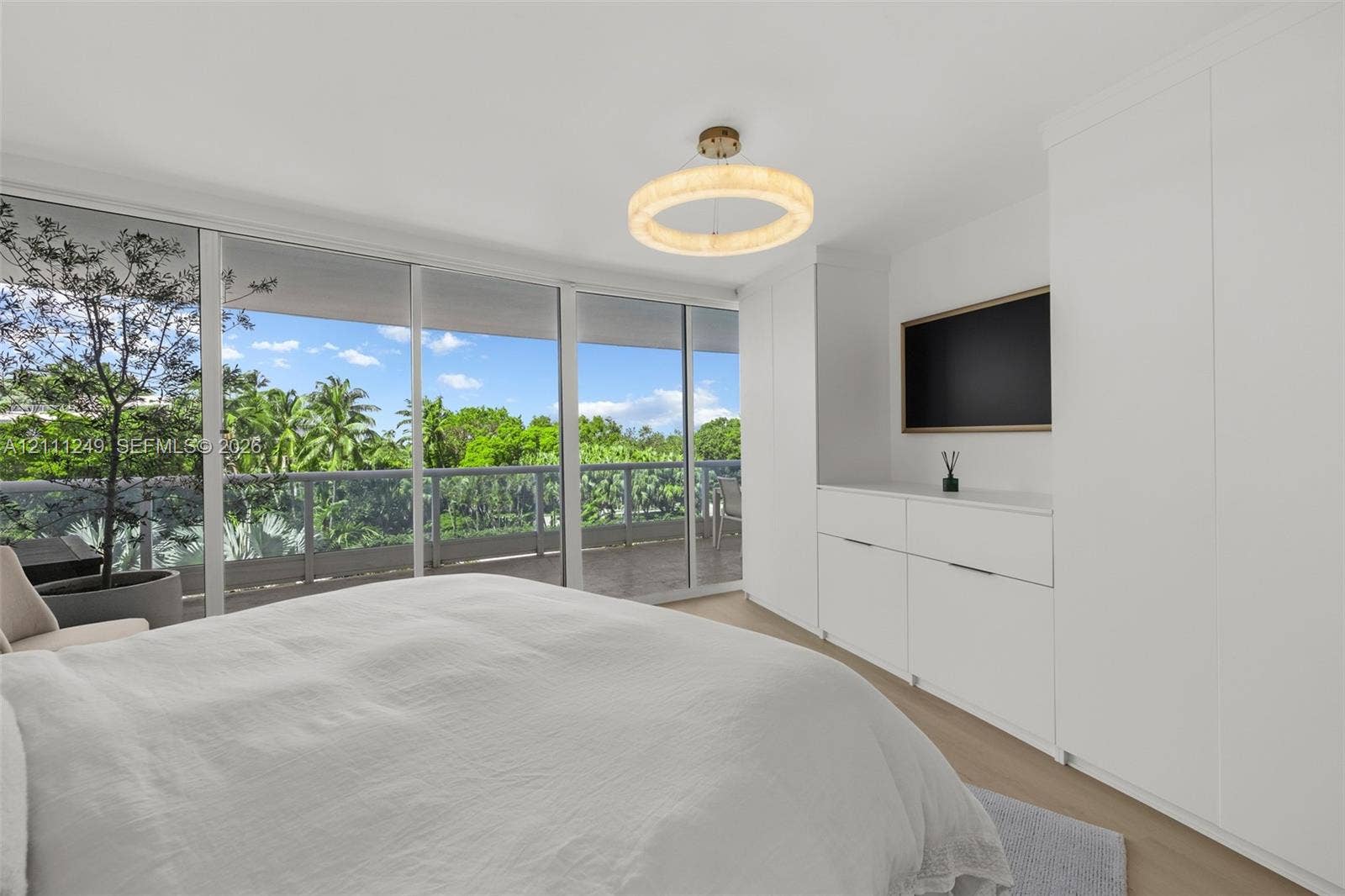 Floor-to-ceiling glass sliding doors, built-in floor-to-ceiling white cabinetry, circular textured flush-mount light fixture, light wood-finish flooring, and integrated balcony with metal railing