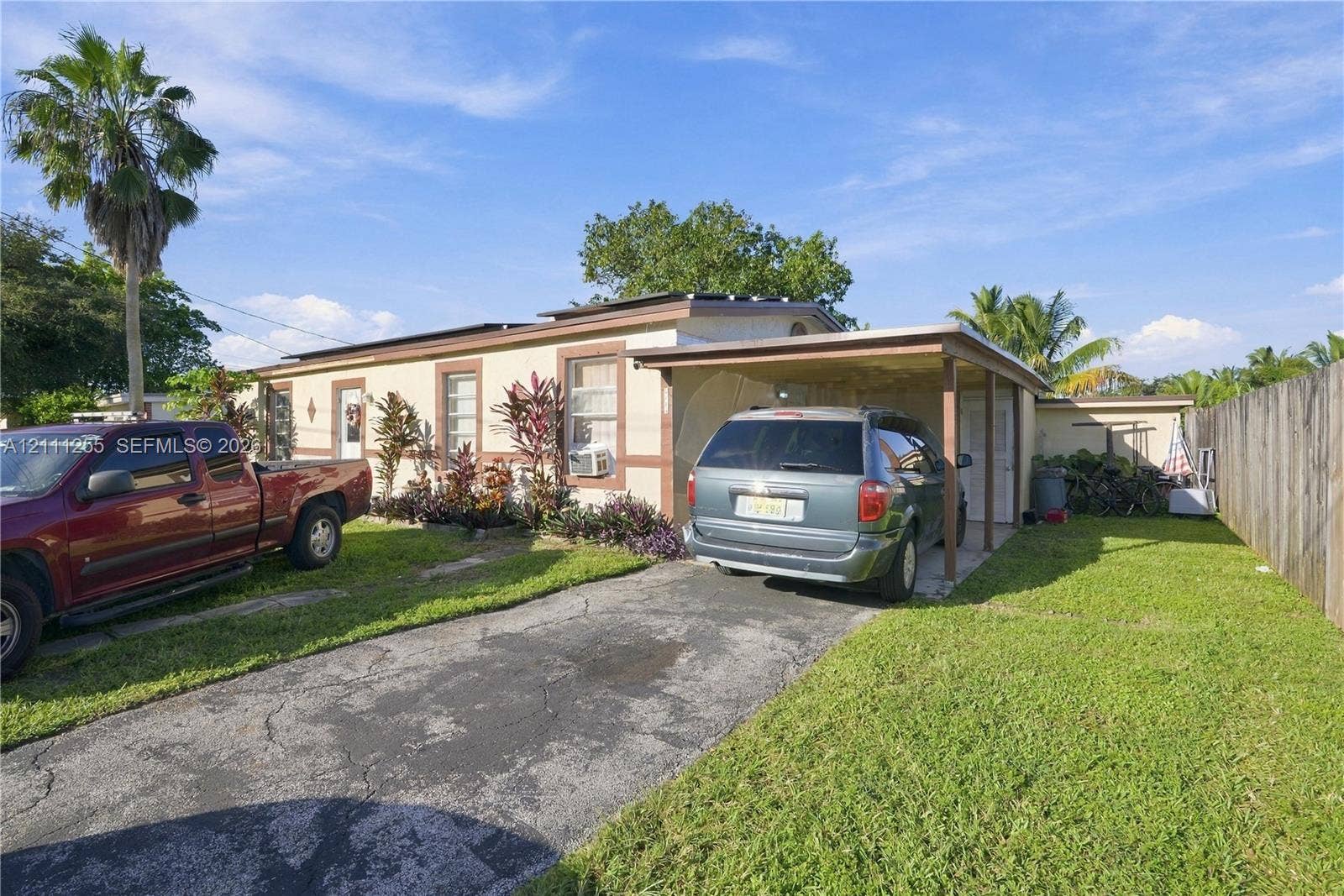 Single-story exterior featuring a paved driveway, attached carport, perimeter wood fencing, and mature palm landscaping
