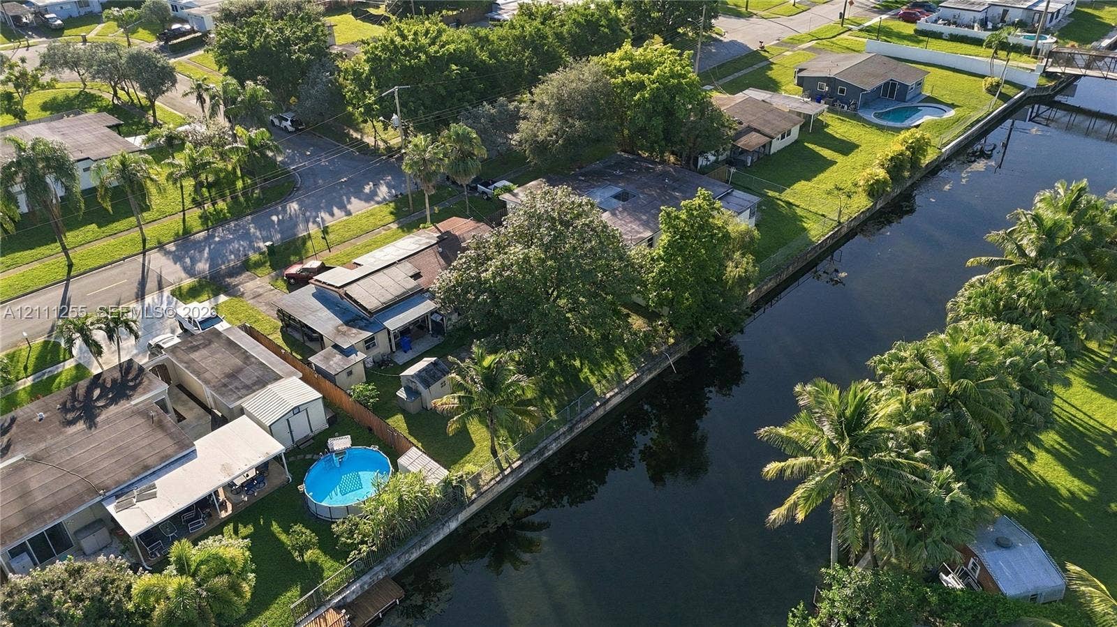 Canal-front property featuring a detached swimming pool, mature palms, and fenced perimeter