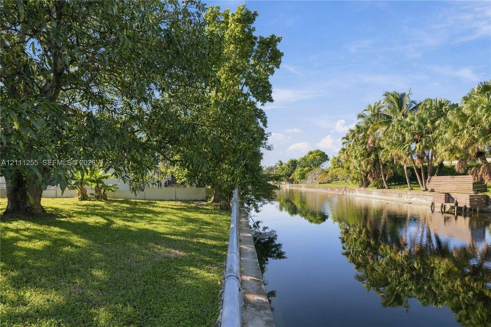 Waterfront lot featuring a concrete bulkhead, mature trees, manicured lawn, and perimeter fencing