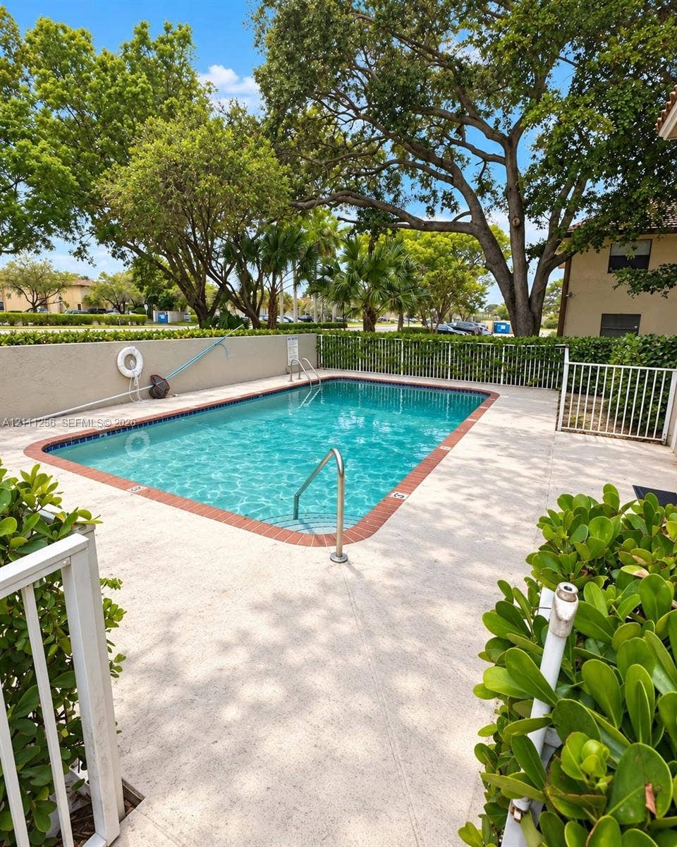 Rectangular swimming pool featuring a brick-tiled border, metal handrail, concrete patio surround, and white perimeter fencing