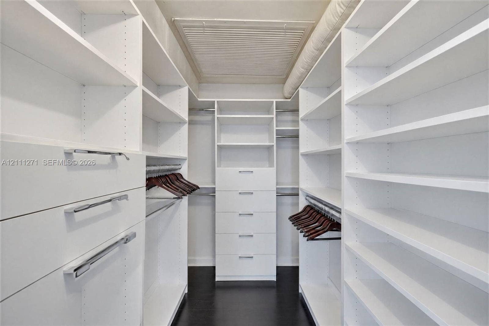 Walk-in closet featuring extensive built-in white shelving, multiple storage drawers with silver-tone hardware, and wood-finish flooring
