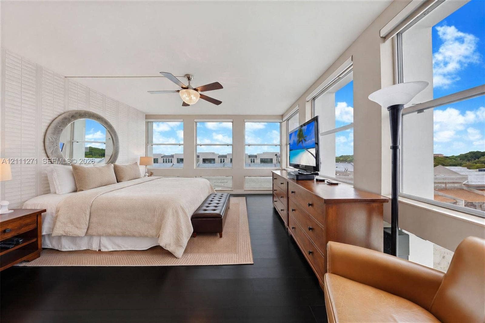 Spacious room featuring floor-to-ceiling windows, dark wood-finish flooring, a wall-mounted circular mirror, and a ceiling fan with integrated lighting