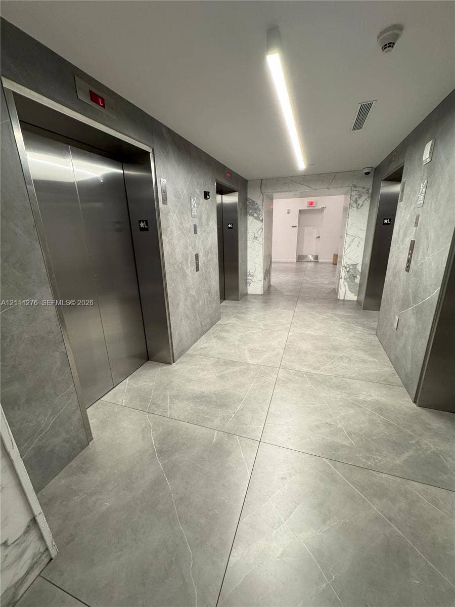 Elevator lobby featuring large-format stone-finish floor and wall tiles, stainless steel elevator doors, linear recessed lighting, and white stone-patterned column detailing