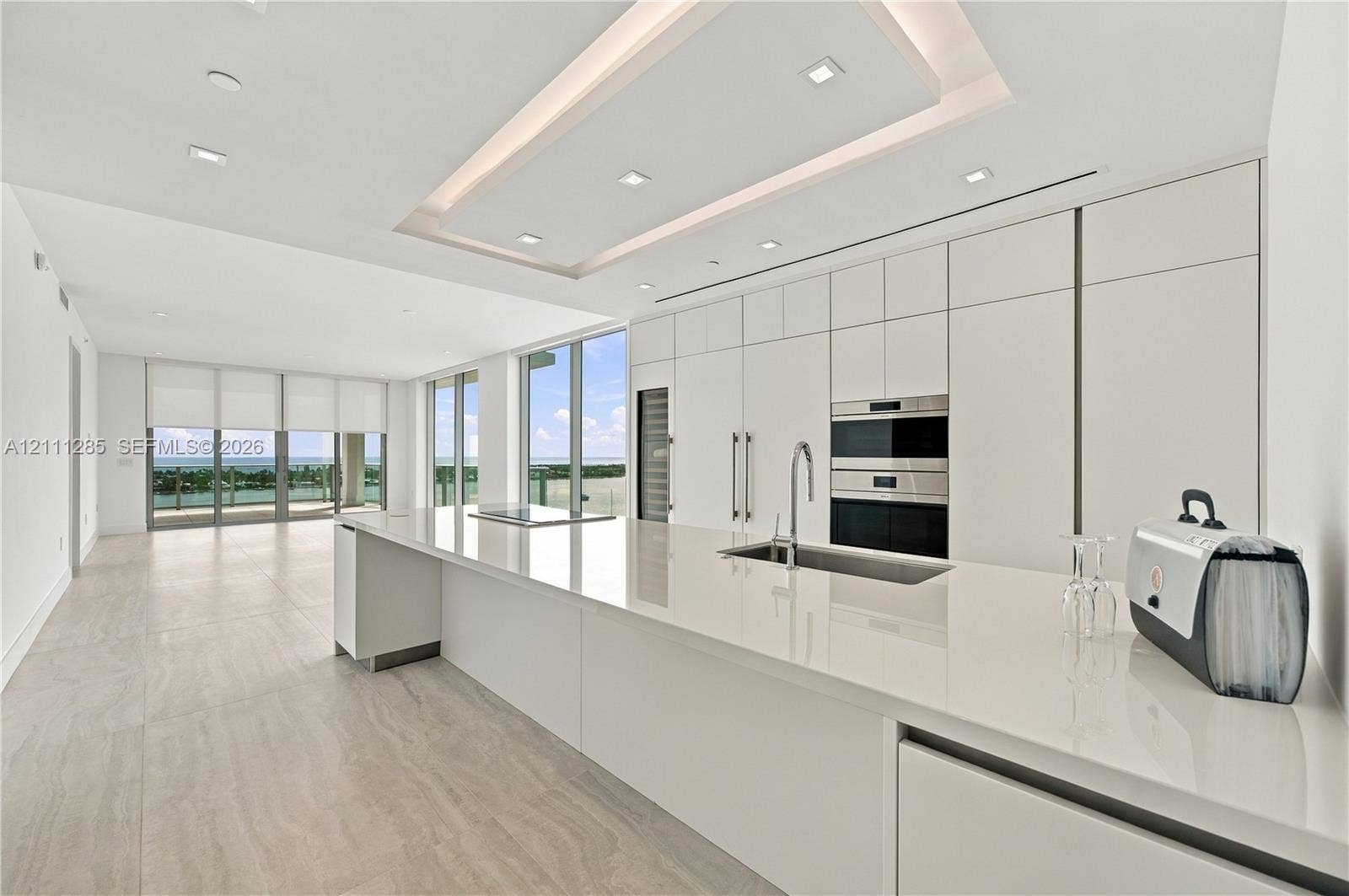 Modern kitchen featuring seamless flat-panel cabinetry, a spacious solid-surface island, built-in stainless steel appliances, recessed tray ceiling lighting, and large-format tile flooring