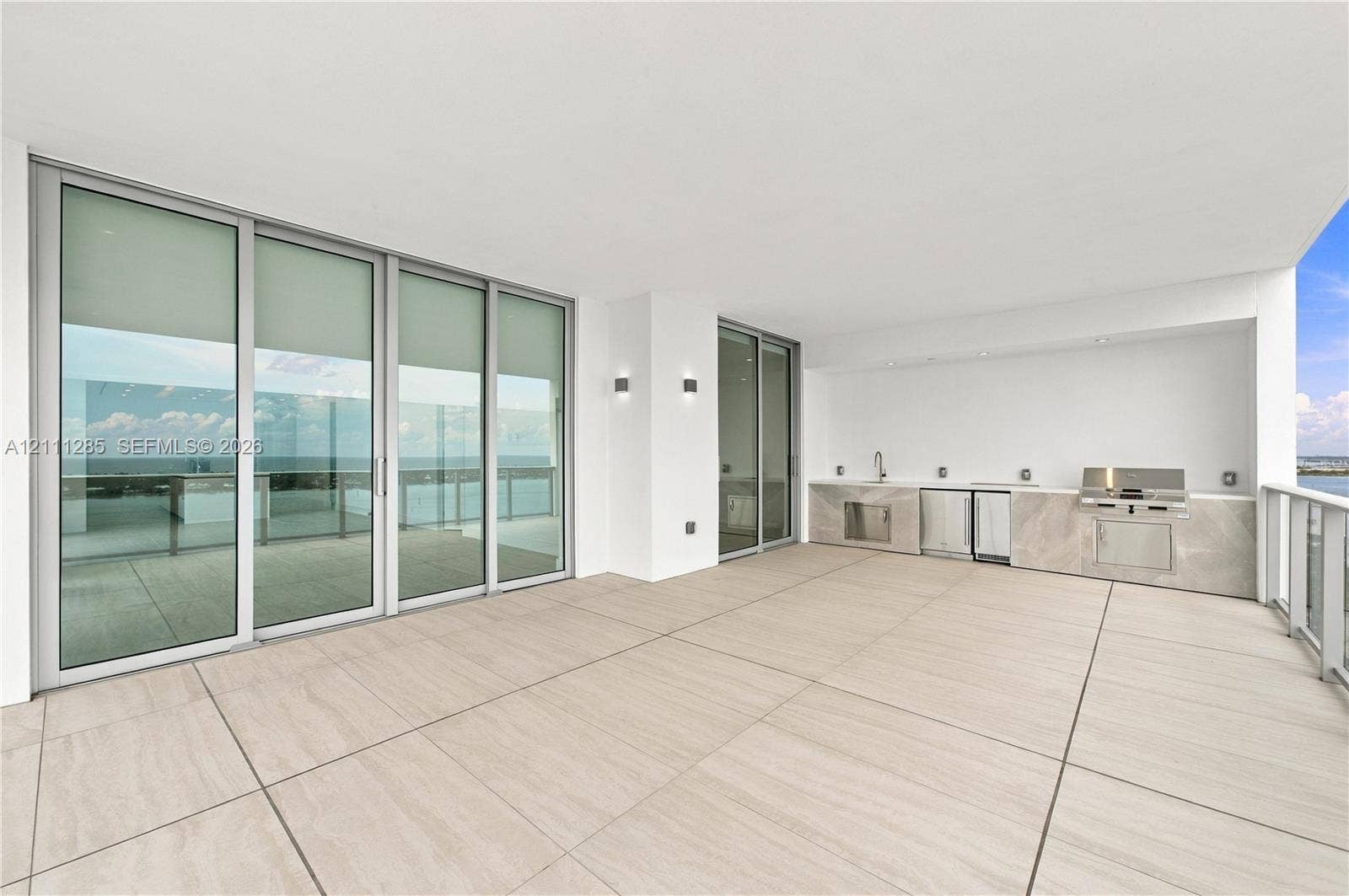 Expansive covered terrace featuring large-format tile flooring, a built-in outdoor kitchen with stainless steel appliances, stone-finish cabinetry, floor-to-ceiling glass sliding doors, and recessed lighting