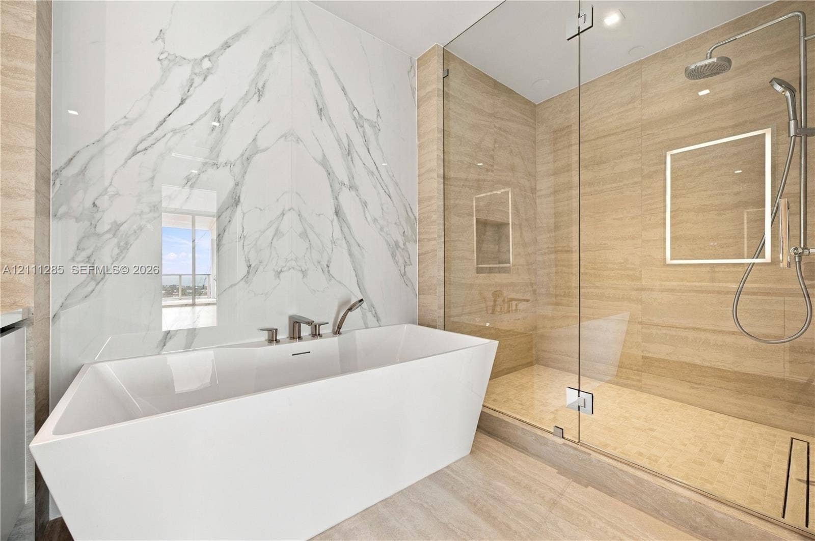 Freestanding soaking tub with a wall-mounted faucet, large-format stone-tile wall, glass-enclosed shower, integrated rainfall showerhead, and recessed lighting