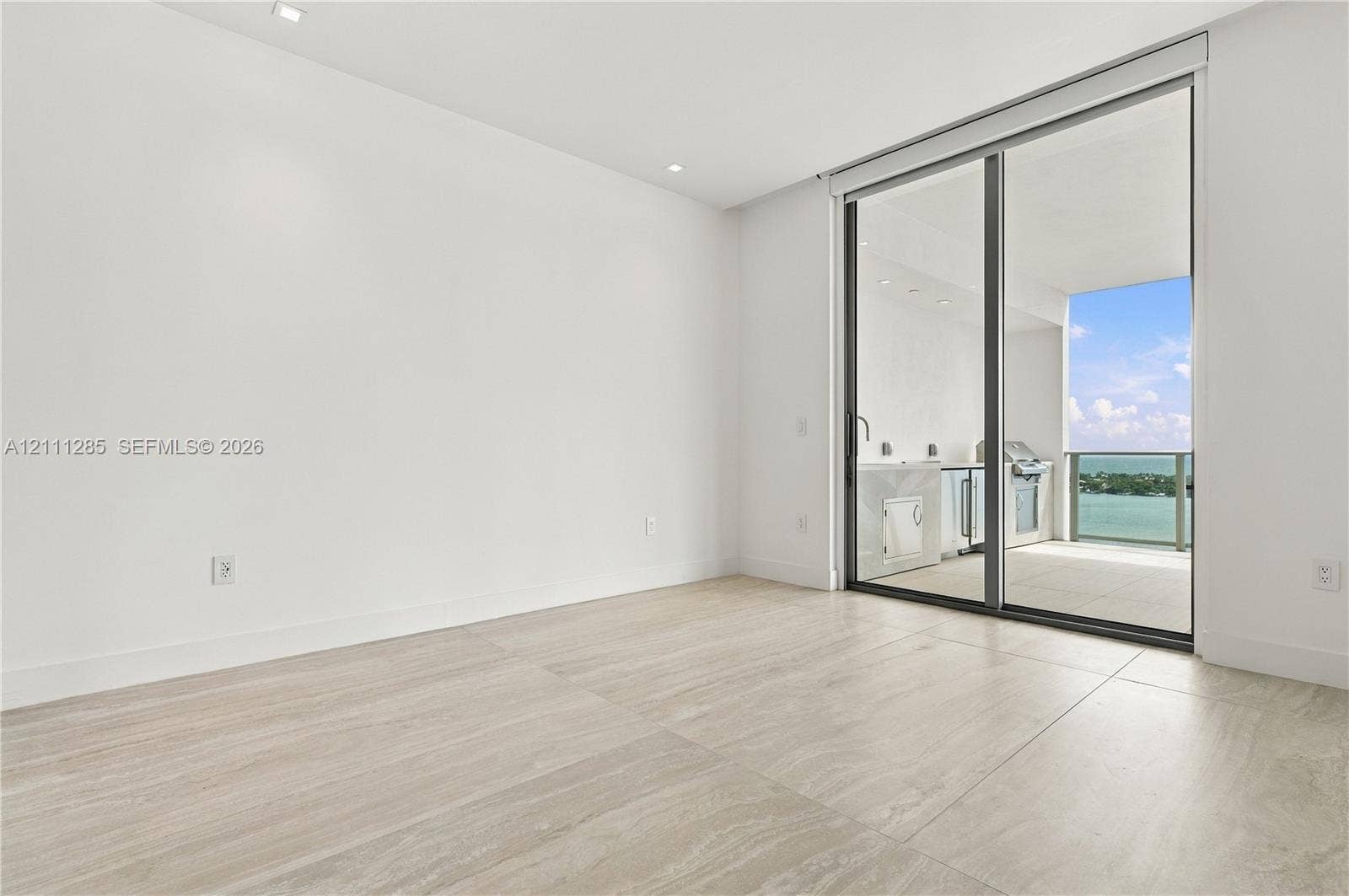 Large-format tile flooring, recessed ceiling lighting, and floor-to-ceiling sliding glass doors opening to a covered balcony with built-in cabinetry