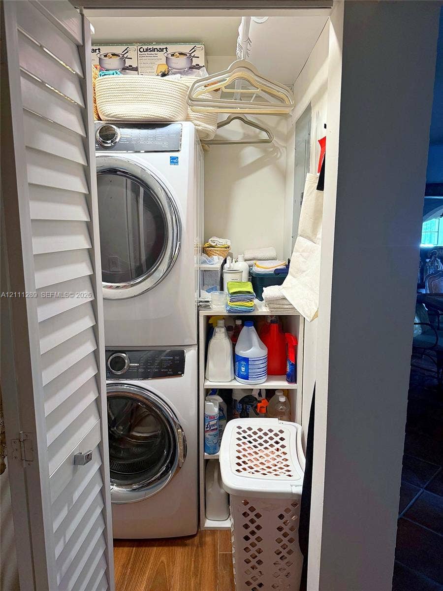 Laundry closet featuring stacked front-load washer and dryer, louvered bi-fold doors, wood-finish flooring, and built-in vertical shelving
