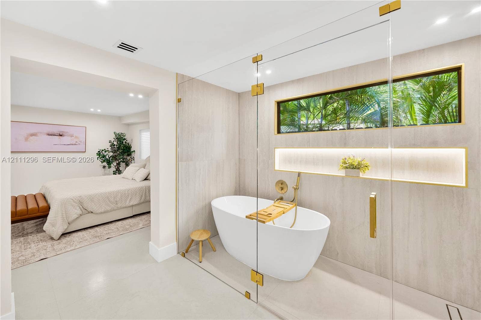 Freestanding white soaking tub enclosed by a frameless glass partition, featuring gold-tone hardware, full-height stone-finish wall tiling, a recessed niche with integrated accent lighting, and a horizontal clerestory window