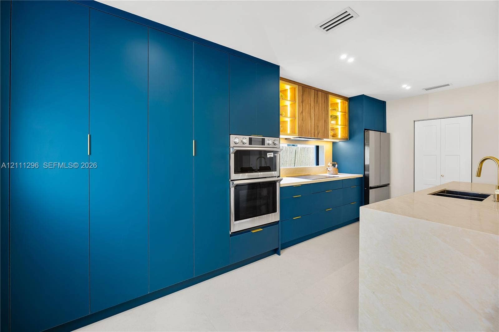 Kitchen featuring floor-to-ceiling blue cabinetry, built-in double wall ovens, wood-finish upper cabinets with integrated lighting, a stone-surfaced kitchen island, and a gold-toned faucet