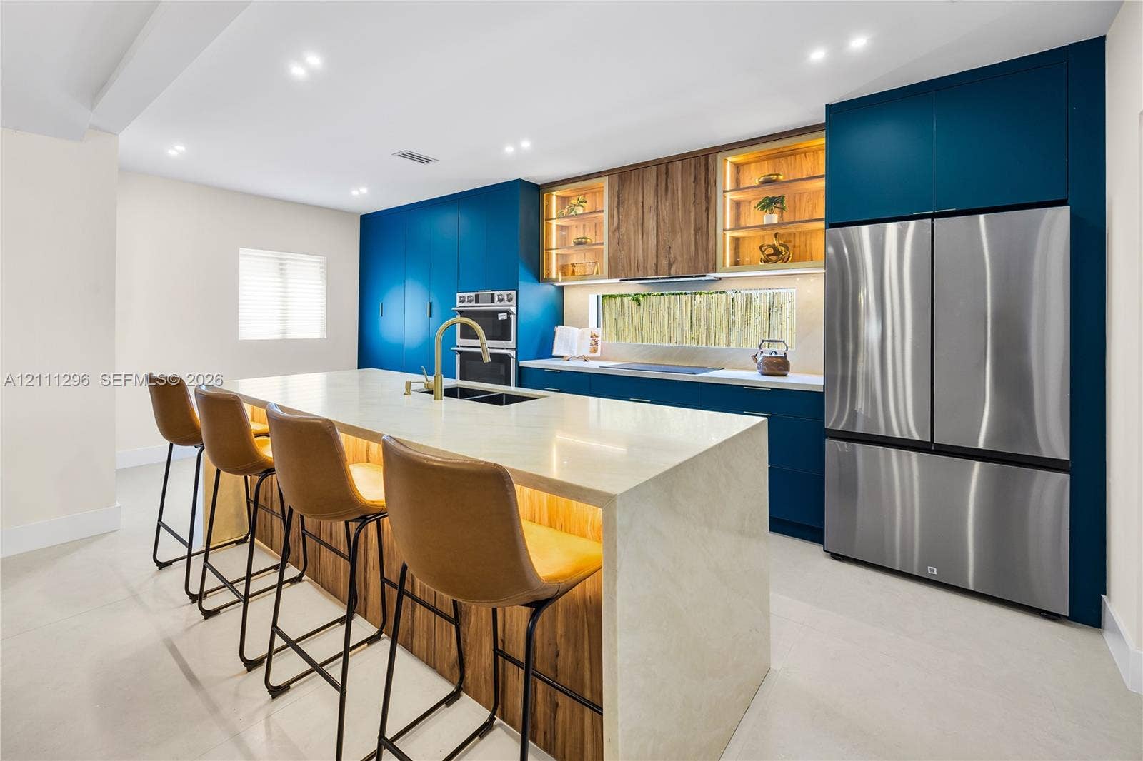 Custom kitchen featuring flat-panel blue cabinetry, expansive stone-surface island with integrated wood-finish base, stainless steel built-in double wall oven, stainless steel refrigerator, and recessed lighting