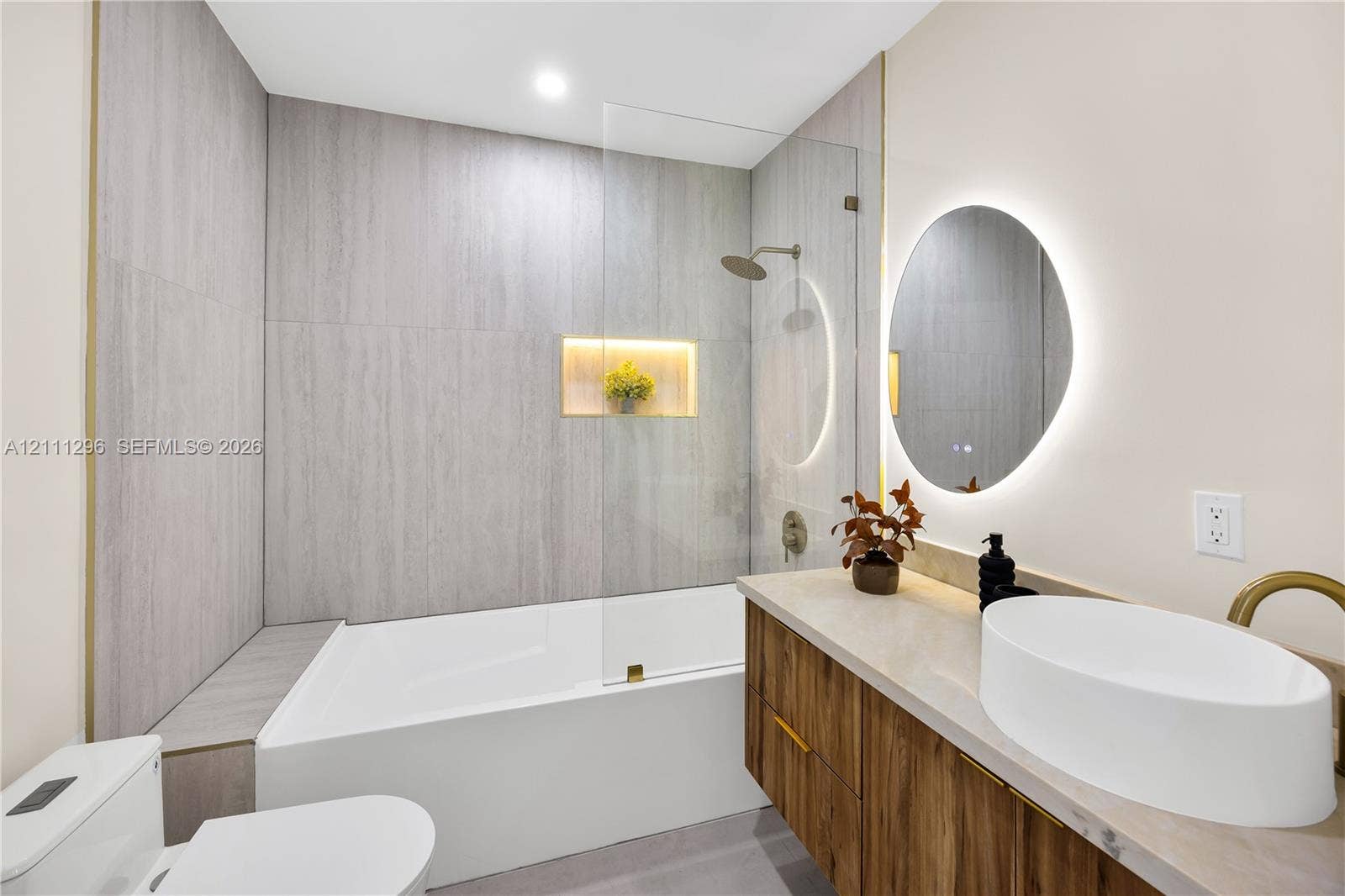 Bathroom featuring a wood-finish vanity with a vessel sink, quartz-style countertop, a walk-in shower with a glass panel and integrated recessed niche, and a circular LED-backlit mirror