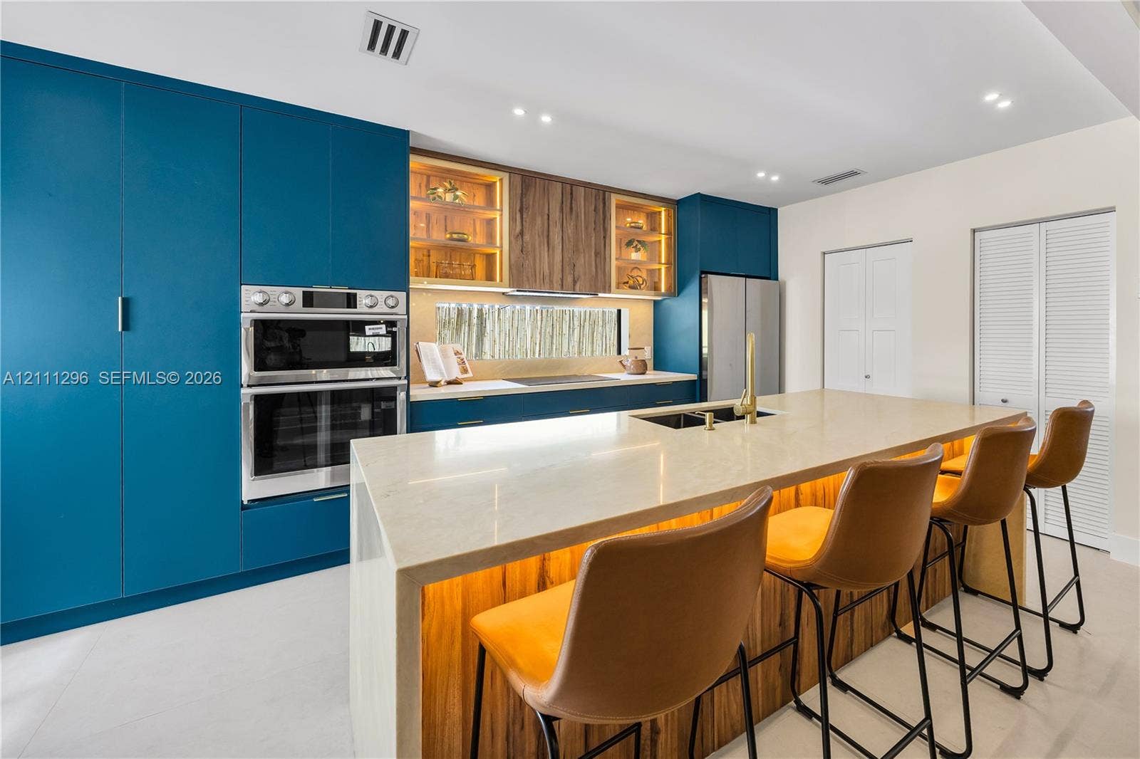 Kitchen featuring matte blue cabinetry, wood-finish upper cabinets with glass shelving, a large island with a stone-surface countertop, and built-in stainless steel double wall ovens