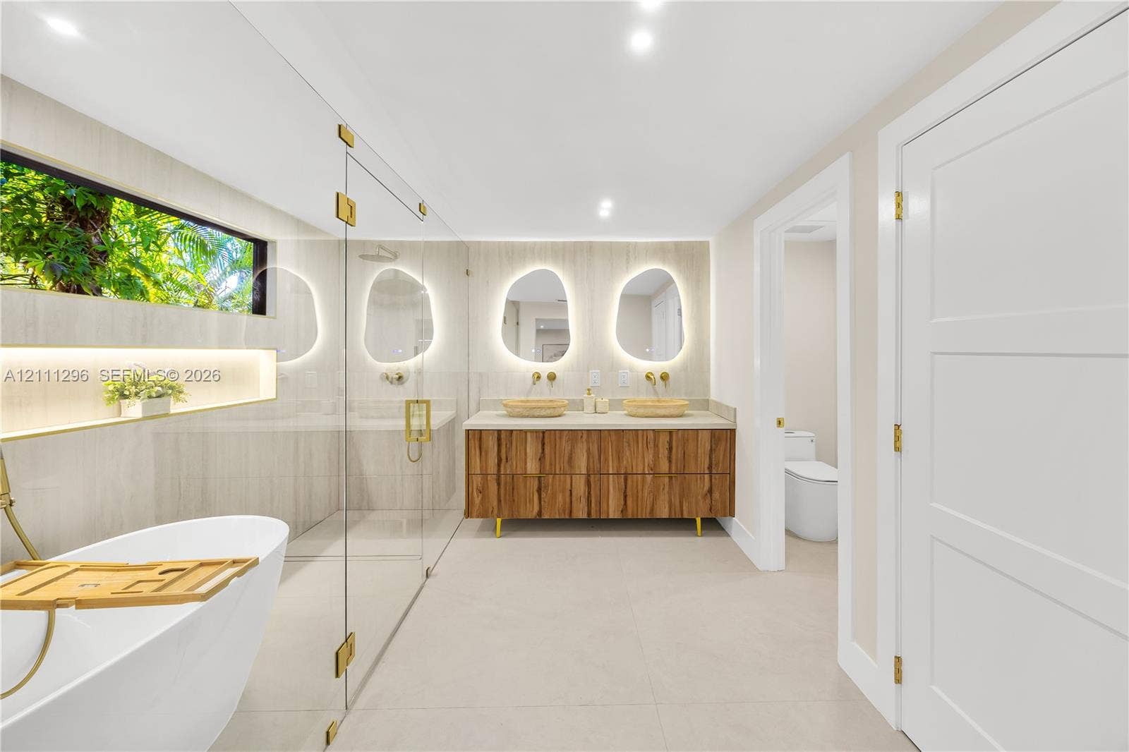 Freestanding soaking tub with gold-tone fixtures, wood-finish floating dual vanity, stone-topped surfaces, wall-mounted faucets, and back-lit organic-shaped mirrors