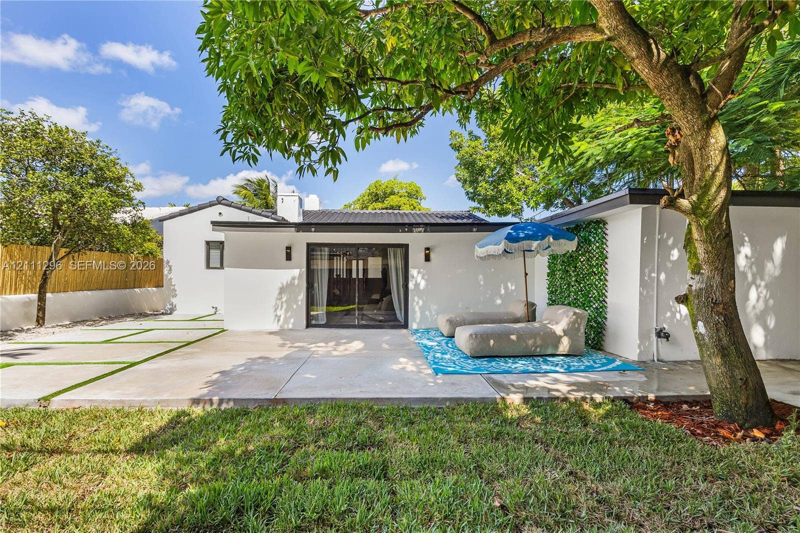 Rear exterior featuring a concrete patio, sliding glass doors, mature trees, a bamboo privacy screen, and exterior wall-mounted lighting