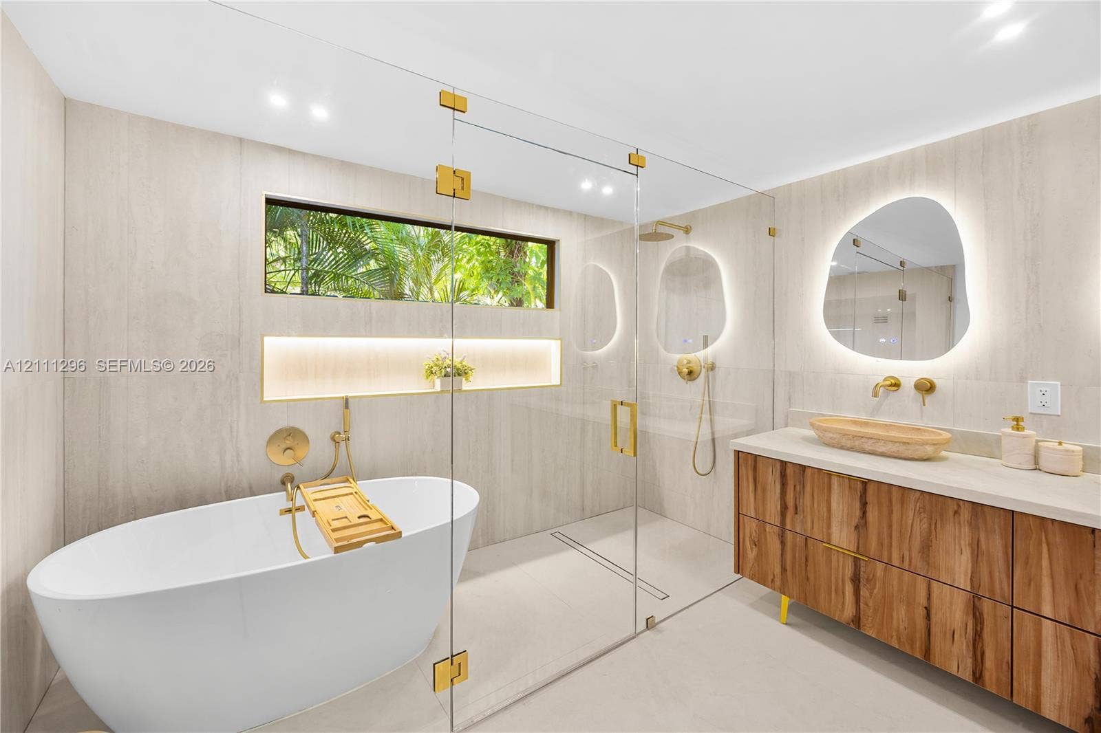 Freestanding soaking tub with gold-tone fixtures, walk-in glass-enclosed shower, wall-to-wall stone-style tile, wood-finish floating vanity with vessel sink, and organic-shaped backlit mirrors