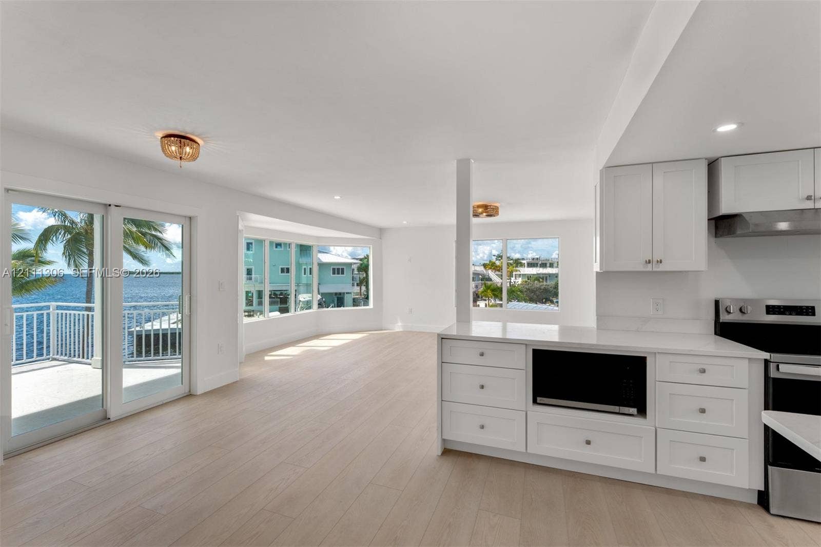 Open-concept kitchen and living area featuring wood-finish flooring, white shaker cabinetry, stainless steel appliances, large bay windows, and sliding glass doors to a waterfront balcony