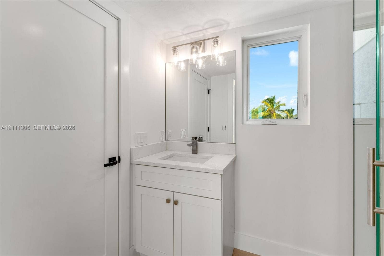 White shaker-style vanity with a solid surface countertop, integrated rectangular sink, three-light fixture with glass shades, wall-mounted mirror, and glass-enclosed shower