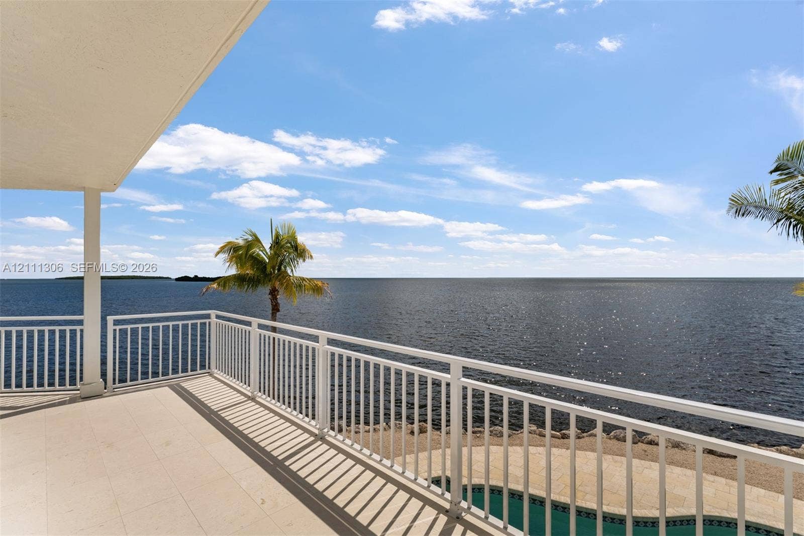 Covered balcony featuring tile flooring and white metal railing, overlooking a waterfront expanse with a pool and palm tree