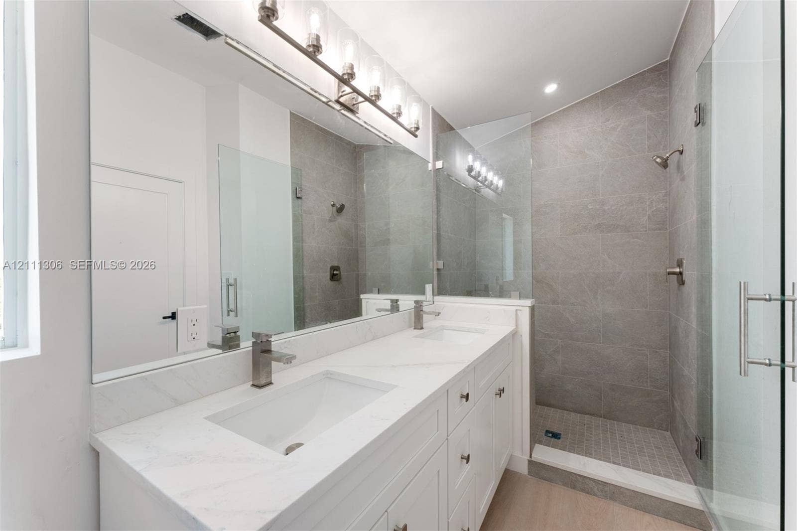 Double-vanity with stone countertop, walk-in shower featuring tile surround and glass enclosure, large wall-mounted mirror, and multi-bulb vanity lighting