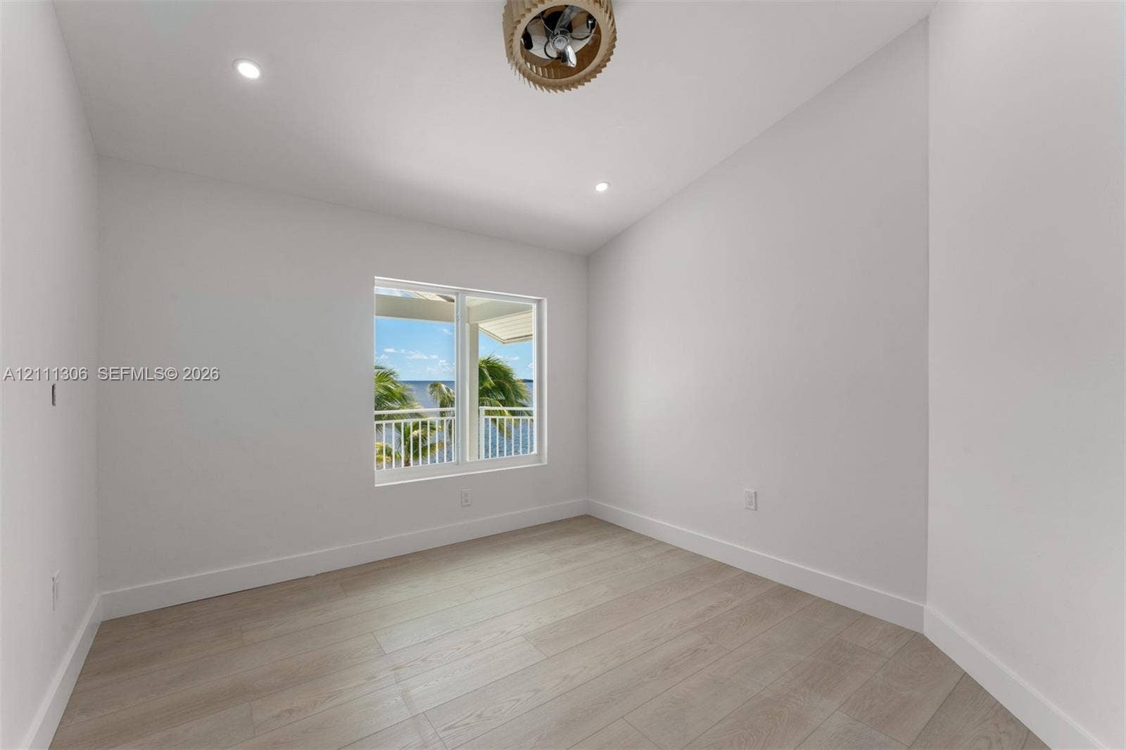 Neutral interior space featuring wood-finish flooring, recessed lighting, and a drum-style ceiling fan, with a window framing water and palm tree scenery