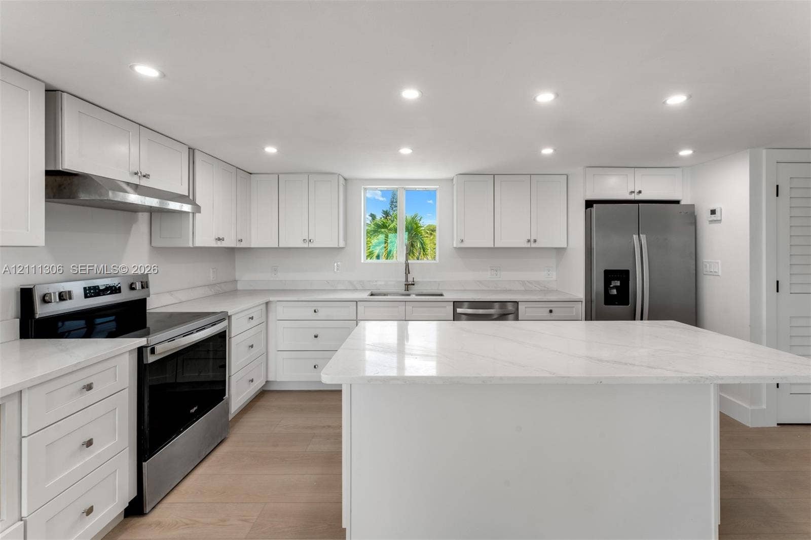 Kitchen featuring white shaker-style cabinetry, a stone-finish island with waterfall edge, stainless steel appliances, recessed ceiling lighting, and light wood-finish flooring