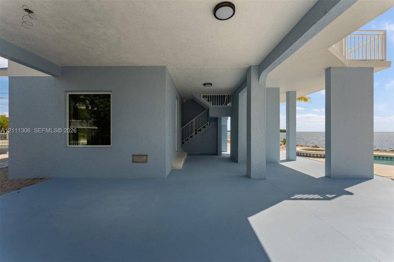 Covered ground-level patio featuring structural concrete columns, staircase with metal railings, flush-mount lighting, and exterior access to a pool area with water backdrop
