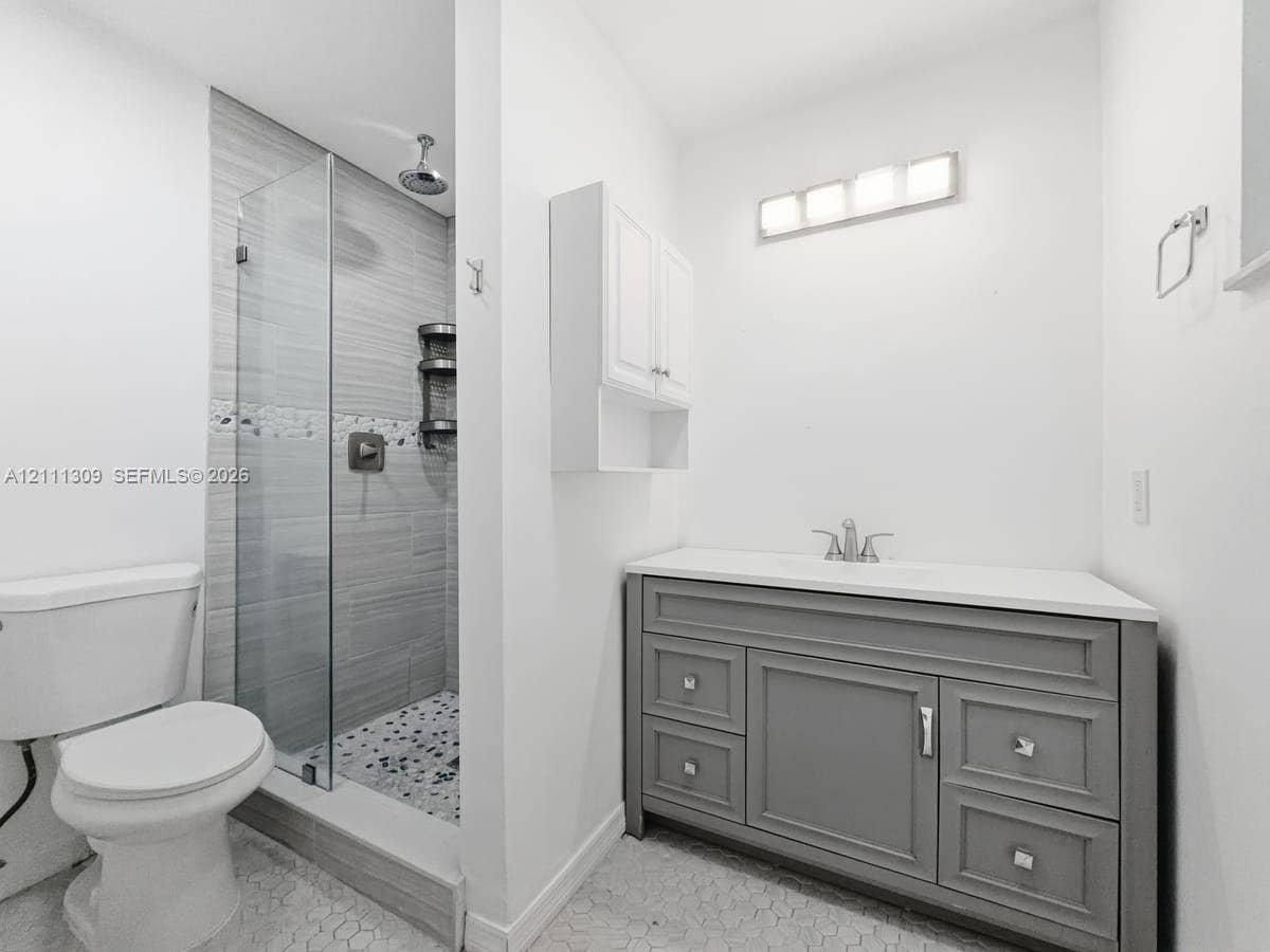 6518 Kendale Lakes Drive, #809, Miami, Miami, FL 33183 Bathroom featuring a grey vanity with a solid surface countertop, glass-enclosed walk-in shower with horizontal tile walls, pebble-style floor tiling, and wall-mounted cabinetry
