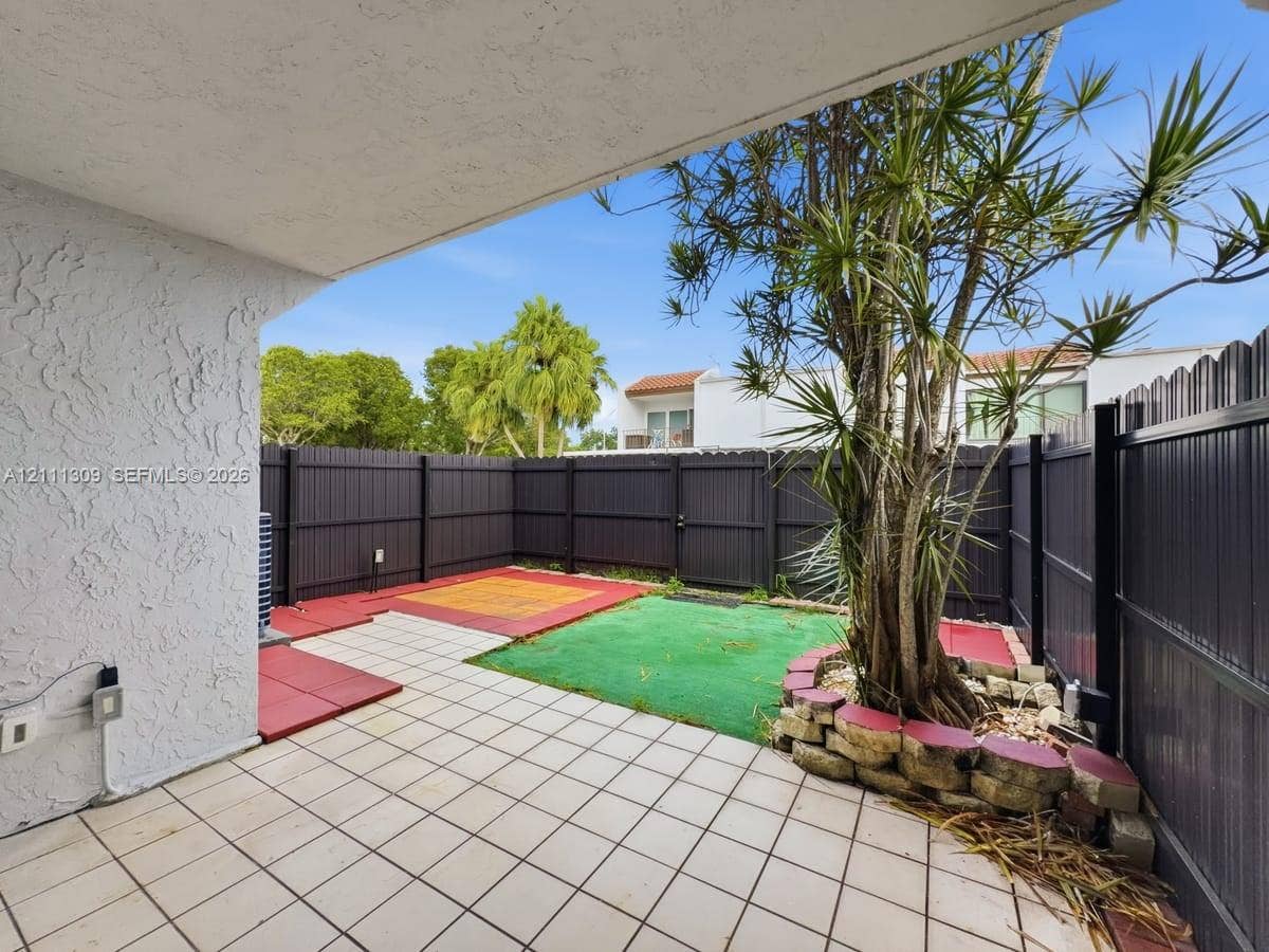 6518 Kendale Lakes Drive, #809, Miami, Miami, FL 33183 Covered patio with tiled flooring, enclosed by dark vertical-panel fencing, featuring a raised stone planter border and mature landscaping